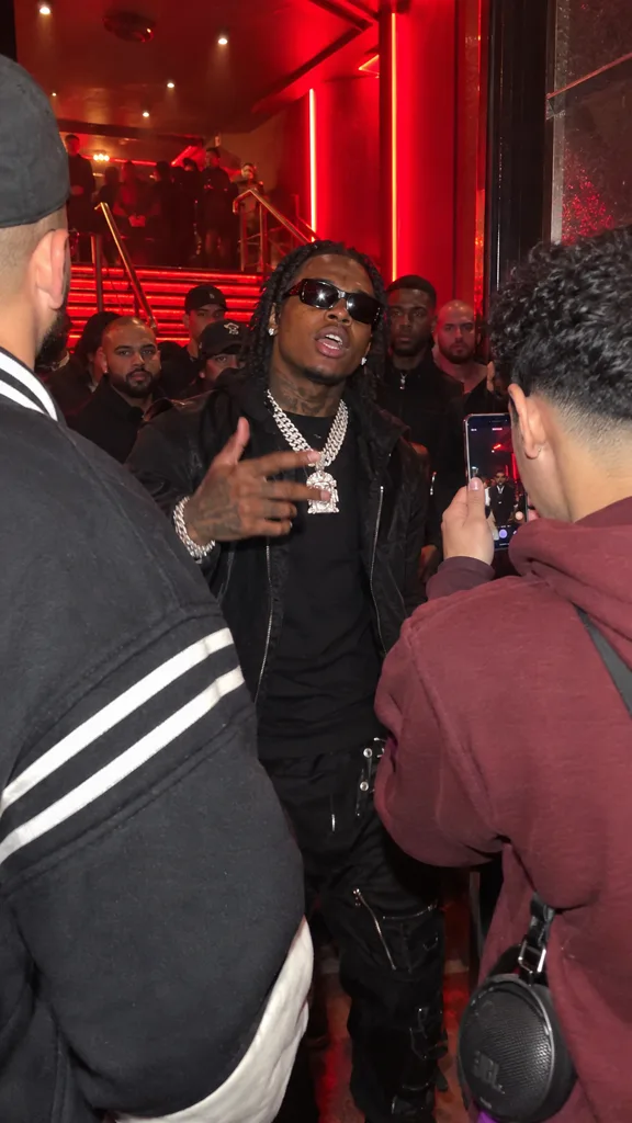 A rapper in sunglasses and a chain freestyling while friends film on their phones